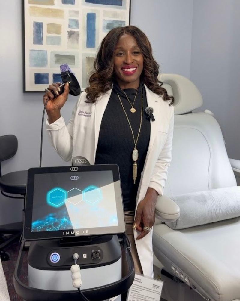 Dr. Kerline Marcelin, MD, smiling in a Hudson Aesthetics MD treatment room, holding the Morpheus8 handpiece beside the InMode platform